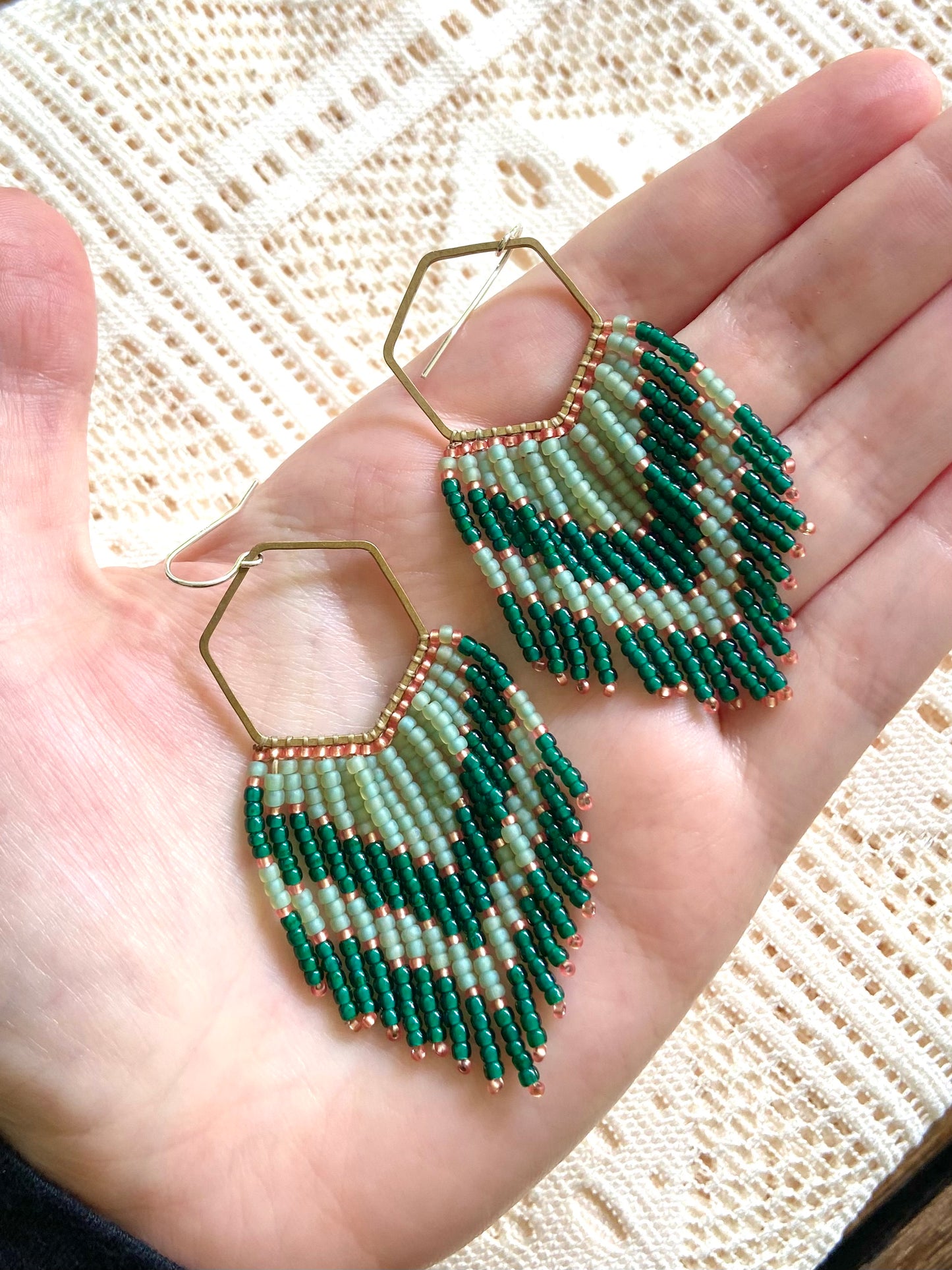 Green beaded earrings held in a hand against a textured background