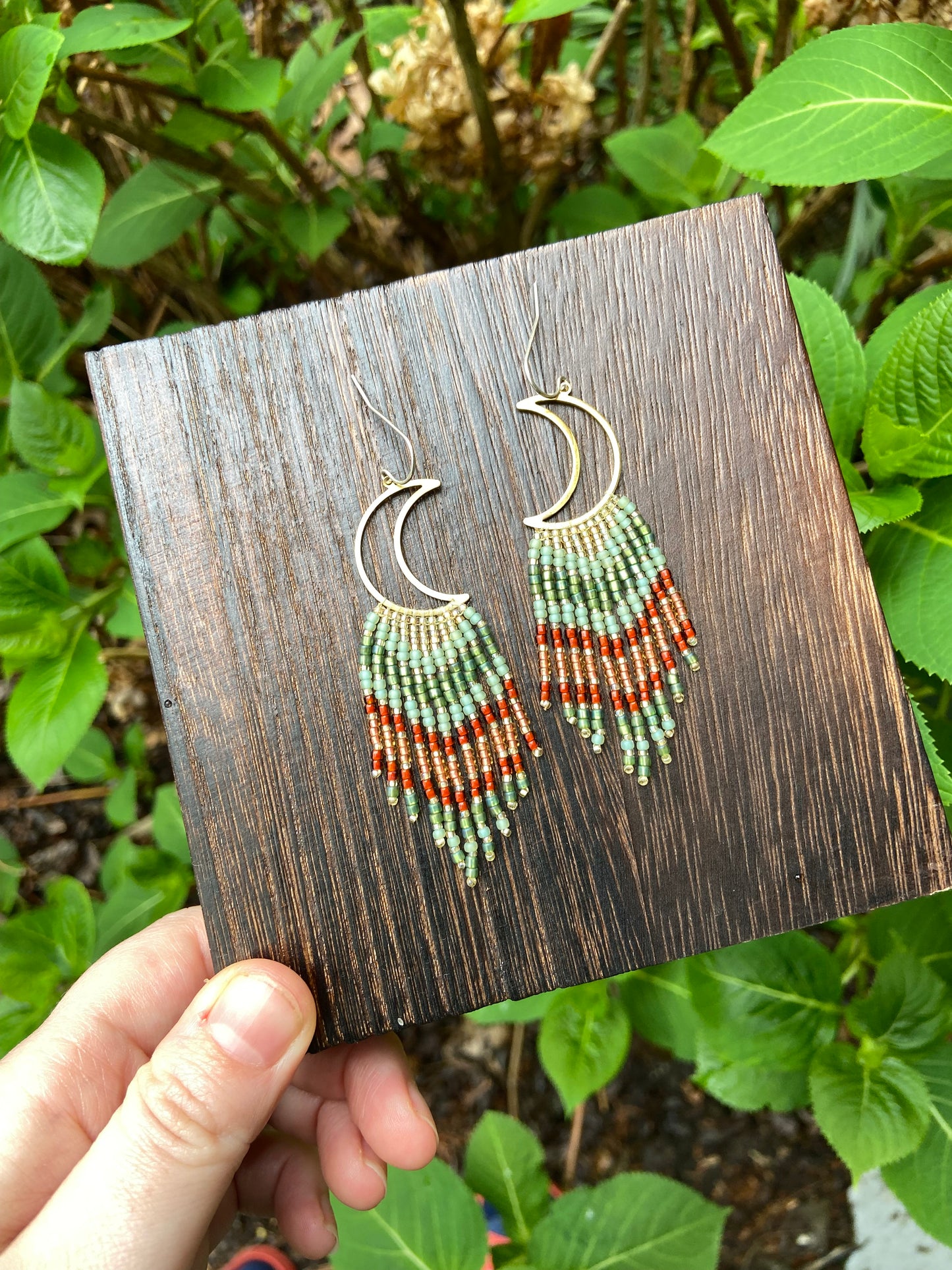 Hand holding a wooden display card with beaded earrings against a natural background