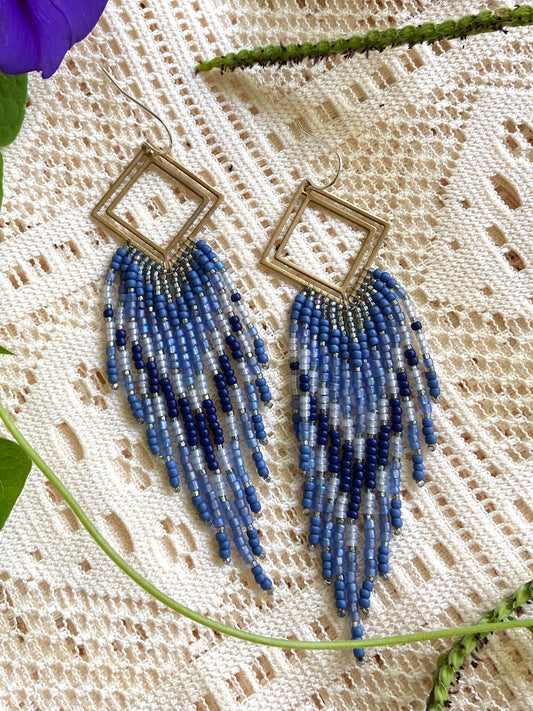 Blue beaded earrings on a textured fabric background with a purple flower and green leaves.